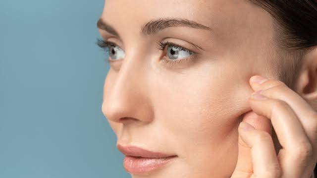 Editorial close-up of a woman's face in profile with a hand gently touching the cheek, highlighting firm, smooth skin against a soft blue backdrop